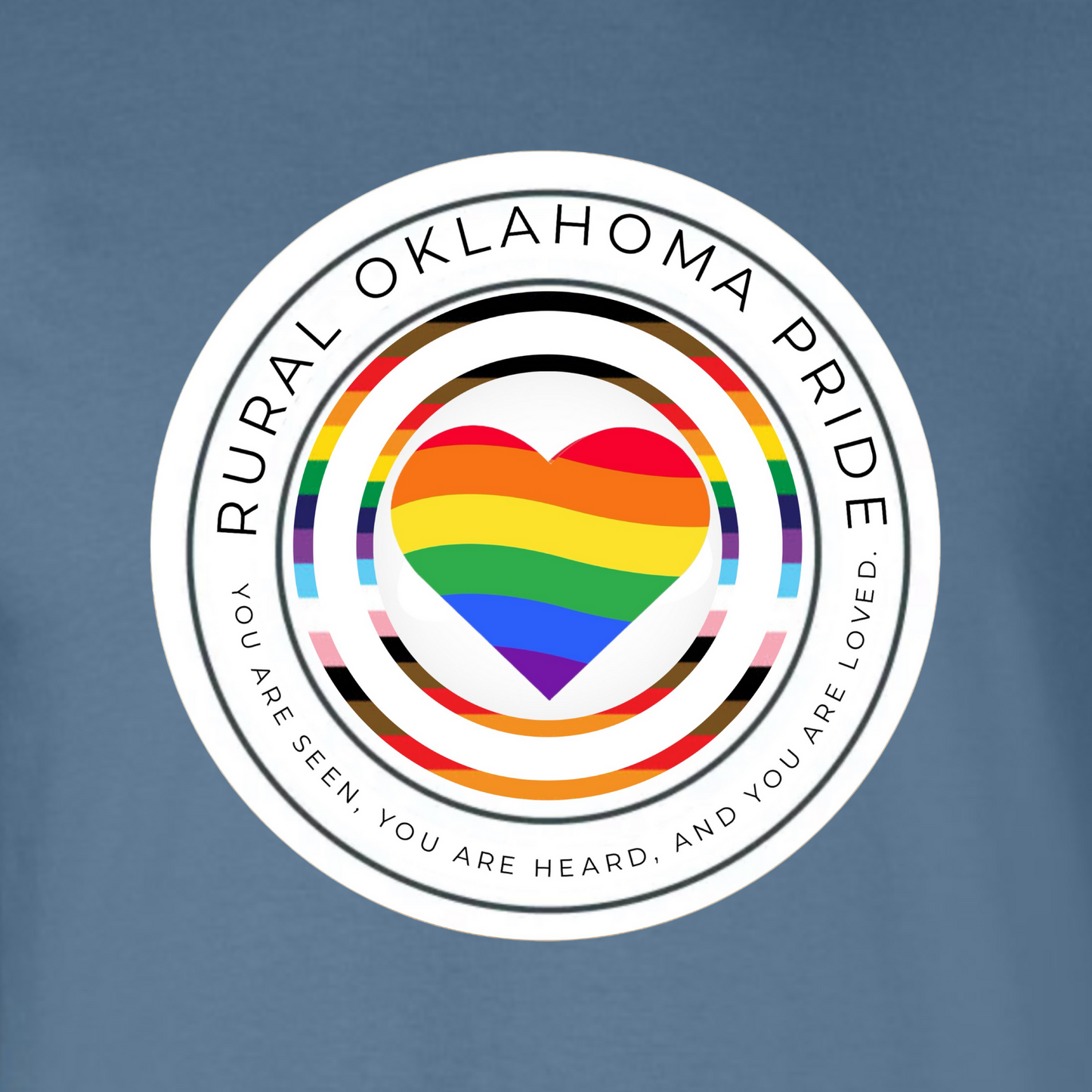 On an indigo blue background is the rural oklahoma pride logo. The logo has Rural Oklahoma Pride written along the top half of a white circle. The bottom half reads You are seen You are Heard and You are Loved there is the outline of another circle the colors of the Daniel Qasar pride flag with an identical outline slightly smaller inside of it. At the very center of the design is a heart that is colored with waves of color that form the rainbow. 